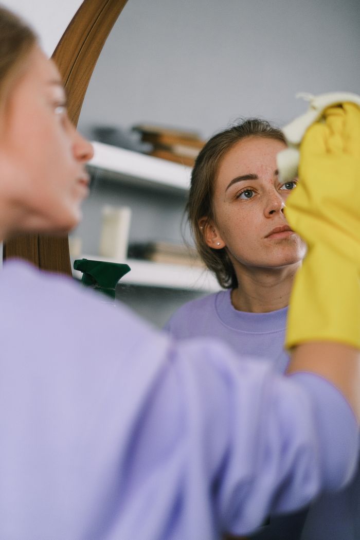 home-img Crop female in purple sweatshirt and latex gloves washing mirror in light apartment in daytime